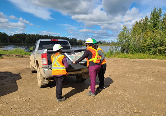 Three people standing at the back of a utility truck near a body of water looking at a map.