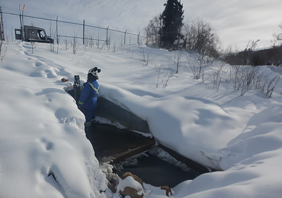 A person in protective clothing standing in a hole in the snow taking measurements.